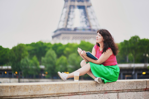 Girl with coffee to go reading a book near the Eiffel tower. - Фото, изображение