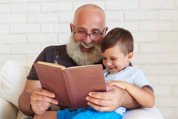 Aged man with white beard reading a book to his grandson at home - Valokuva, kuva