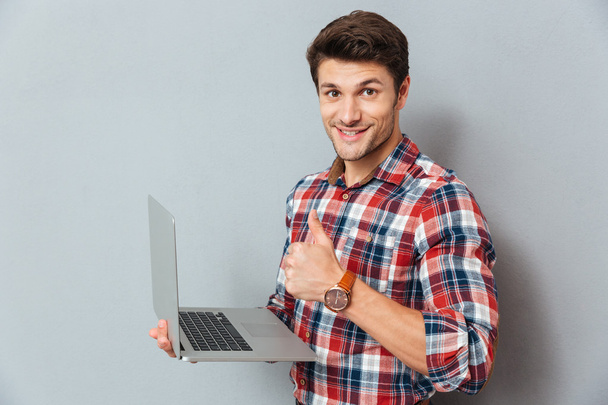 Cheerful young man holding laptop and showing thumbs up - Foto, Imagem