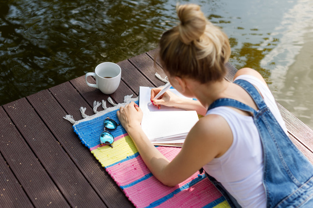 Woman making plans, taking notes in calendar or writing in her diary lying on the pier and drinking hot coffee. Daydreaming, planning concept - Photo, image