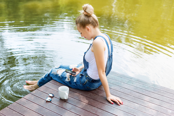 Young woman using her smartphone sitting on the pier and drinking hot coffee - Foto, Bild