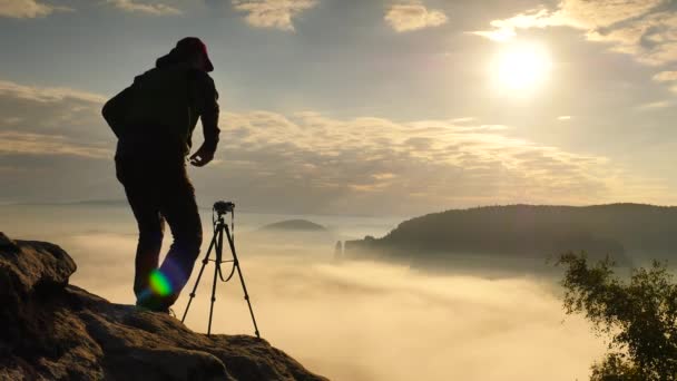 Fotograf w zielonej wiatrówka i czarne Spodnie trekkingowe zatrzymać aparat na statywie na krawędzi klifu. Dreamy fogy krajobraz, niebieski mglisty wschód słońca w pięknej dolinie poniżej - Materiał filmowy, wideo
