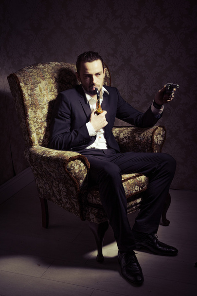 Fiatal jóképű és elegáns férfi ül a székben, kezében pisztoly és dohányzik egy cső elszigetelt felett vintage háttér - Fotó, kép