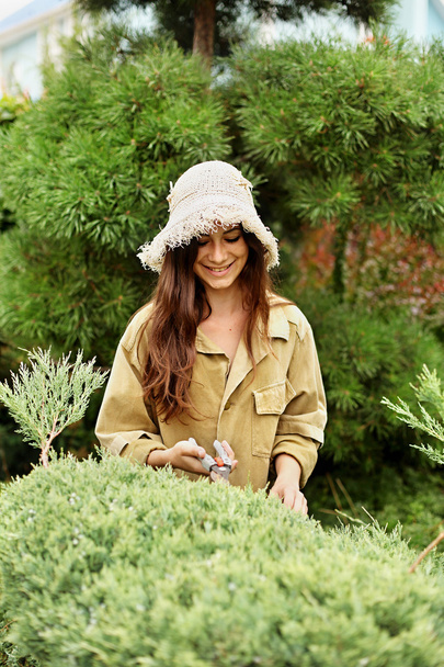 Smiling girl gardener in working clothes and straw hat cuts garden scissors evergreen. - Photo, Image