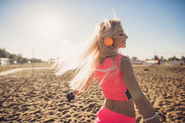 sport outdoor photo of beautiful young blonde woman in pink colorful sport suit listening to music on headphones by the beach - Foto, immagini
