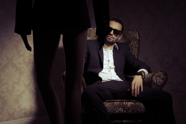 Young and elegant man sitting in chair wearing black suit, white shirt and sunglasses looking at woman standing in front of him isolated over vintage background - Photo, Image