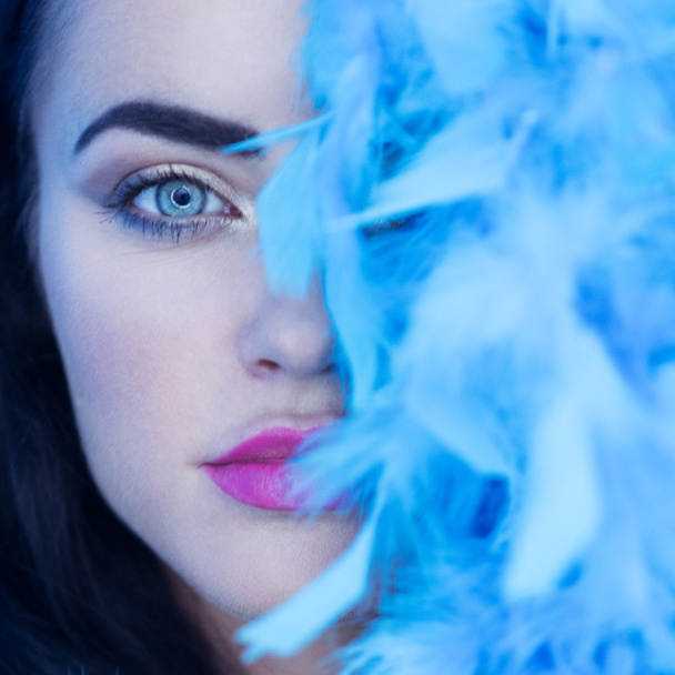 Closeup portrait of beautiful seductive brunette woman smiling and looking into the camera while playing with blue feather boa over blue wall background - Photo, Image