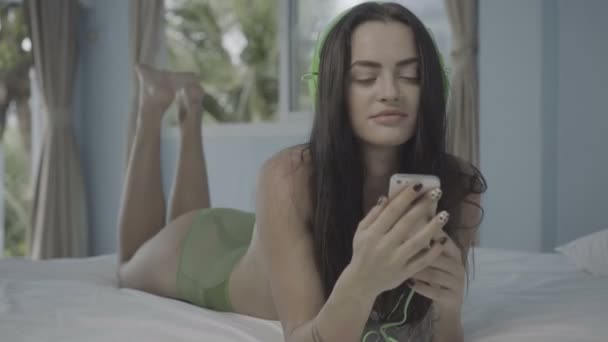 Beautiful young brunette woman lying on bed using her smartphone and headphones - Filmati, video