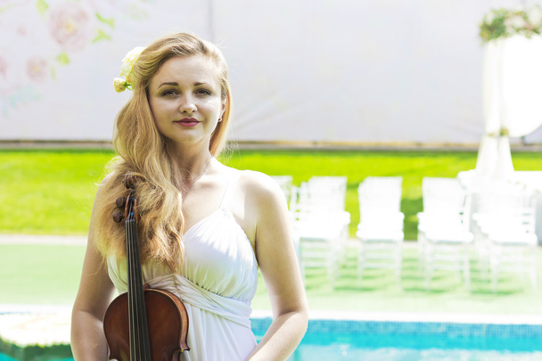 Krásná dívka v bílých šatech na pozadí svatebního oblouku. Dívka s houslemi na pozadí svatebního oblouku. Svatební oblouk poblíž modrého bazénu - Fotografie, Obrázek