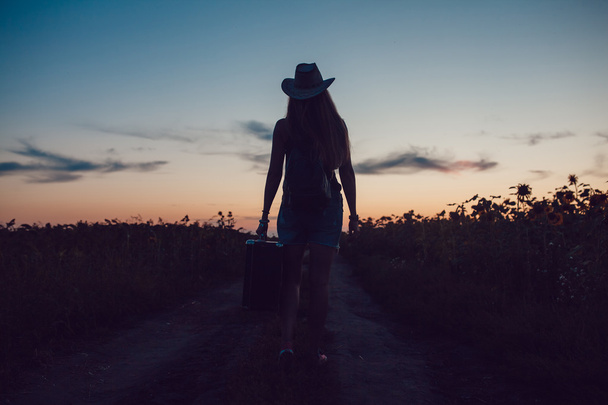Lány egy cowboy kalap állt az úton, a napraforgó mező egy bőrönddel. Várakozás a Súgó. Naplemente. - Fotó, kép