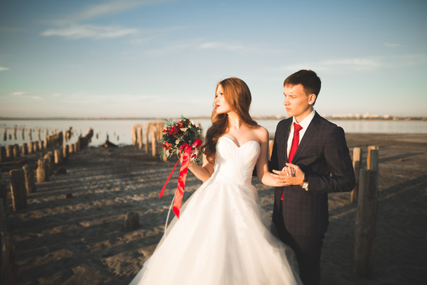 Wedding couple, groom, bride with bouquet posing near sea on sunset - Fotografie, Obrázek