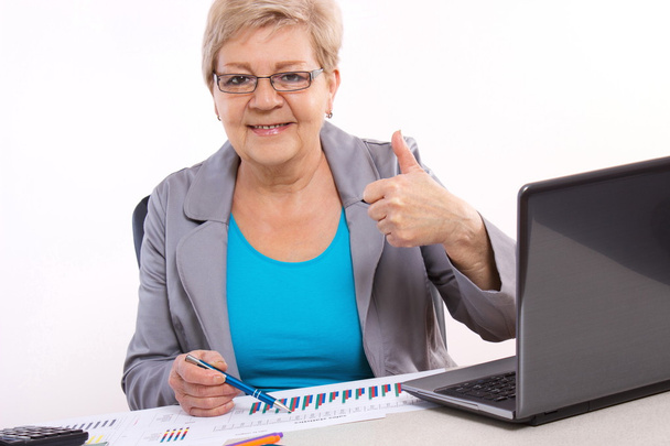 Elderly business woman showing thumbs up and working at her desk in office, business concept - Фото, зображення