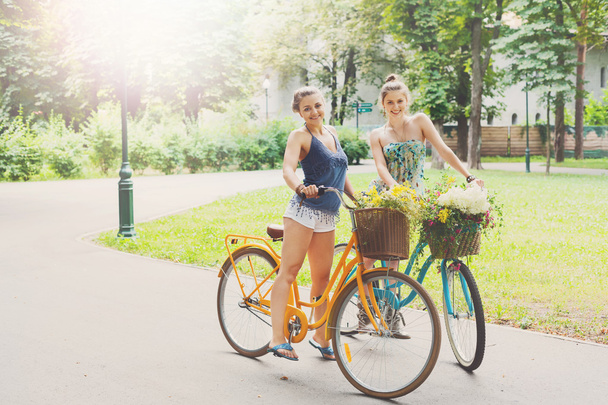 Happy boho chic girls ride together on bicycles in park - Fotó, kép