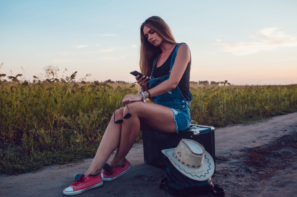 Κορίτσι που κάθεται σε μια βαλίτσα σε ένα ηλιέλαιο πεδίο. Ηλιοβασίλεμα. - Φωτογραφία, εικόνα