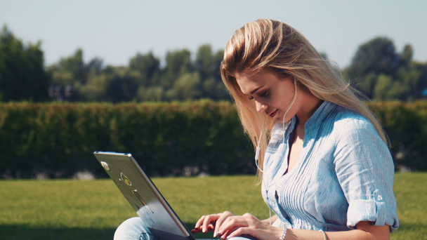 Smiling girl using laptop in earphones on grass - Filmati, video