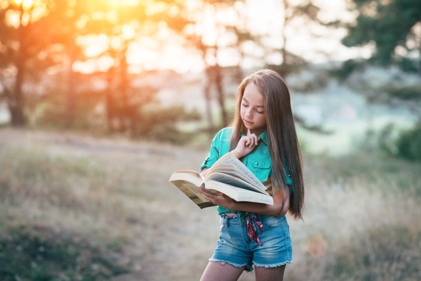 Симпатична дівчинка сидить на траві і читає книгу в
  - Фото, зображення
