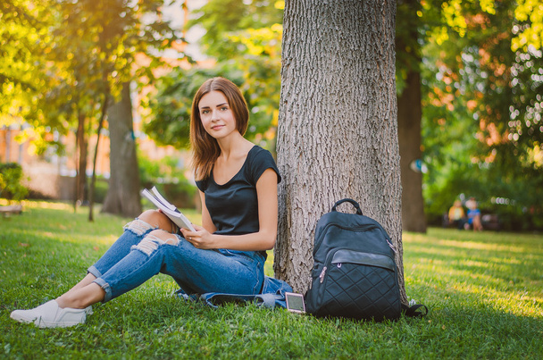 student szczęśliwy dziewczyna siedzi na trawie w pobliżu drzewa, w szczegól - Zdjęcie, obraz