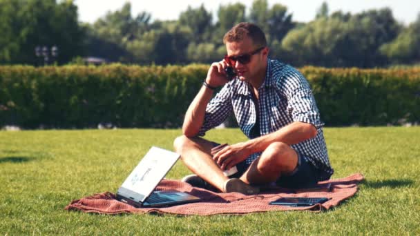 Χαρούμενος νεαρός μιλώντας πάνω από το κινητό τηλέφωνο και να χρησιμοποιούν φορητό υπολογιστή στο πάρκο - Πλάνα, βίντεο