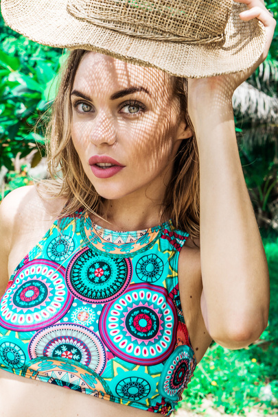 Portrait of beautiful young woman wearing colorful bikini top and holding straw hat in her hand on a sunny day looking into camera over palm trees background. Beautiful eyes closeup of pretty girl with straw hat pattern shadow on her face over green - Fotó, kép