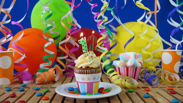 Verjaardag cupcake met kaarsen branden op rustieke houten tafel met achtergrond van kleurrijke ballonnen, plastic bekers en snoepjes met blauwe muur op de achtergrond - Foto, afbeelding