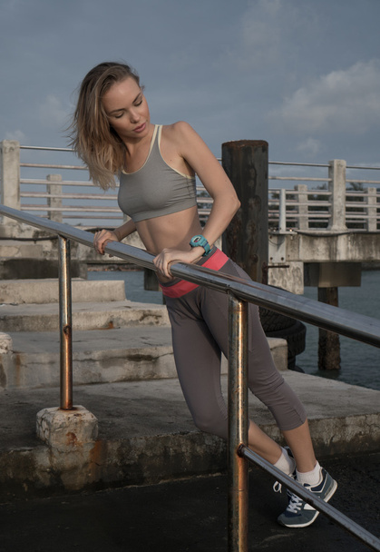 Young pretty blonde female wearing exercise clothing stretching on the pier near the sea and enjoying early morning with beautiful sunrise - Φωτογραφία, εικόνα