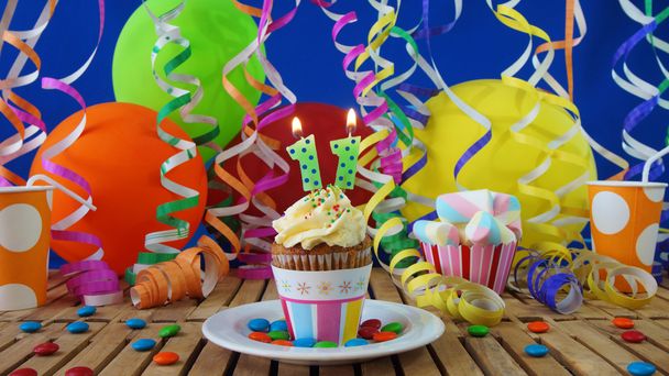 Bolo de aniversário com velas queimando na mesa de madeira rústica com fundo de balões coloridos, copos de plástico e doces com parede azul no fundo
 - Foto, Imagem