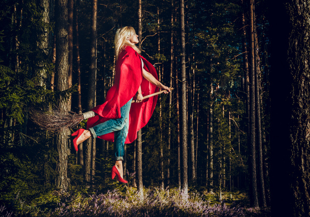 Modern witch blond in jeans and red cape flying on a broom through the forest. Levitation. Halloween holiday. - Photo, Image