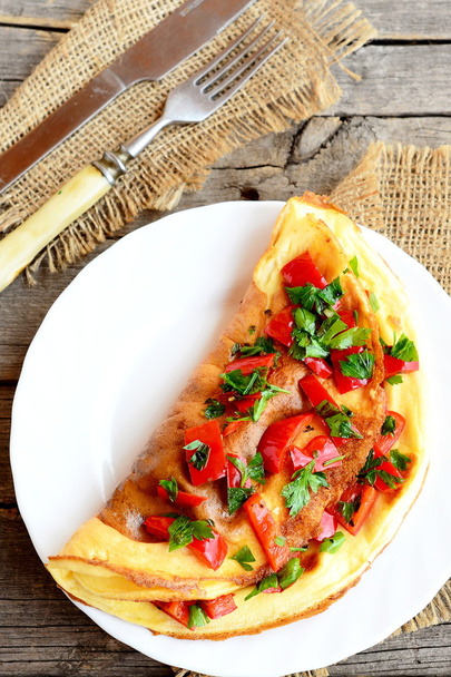 Gevulde ei omelet met groenten op een bord. Vegetarische omelet met gebakken rode peper, verse peterselie en specerijen. Vork, mes op een jute en oude hout achtergrond. Gezond ontbijt recept. Top View  - Foto, afbeelding
