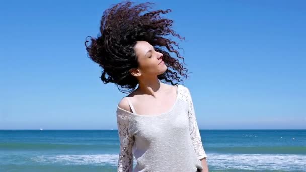 Portret szczęśliwej młodej kobiety potrząsając jej piękne kręcone włosy na brzegu morza, powolny ruch - Materiał filmowy, wideo