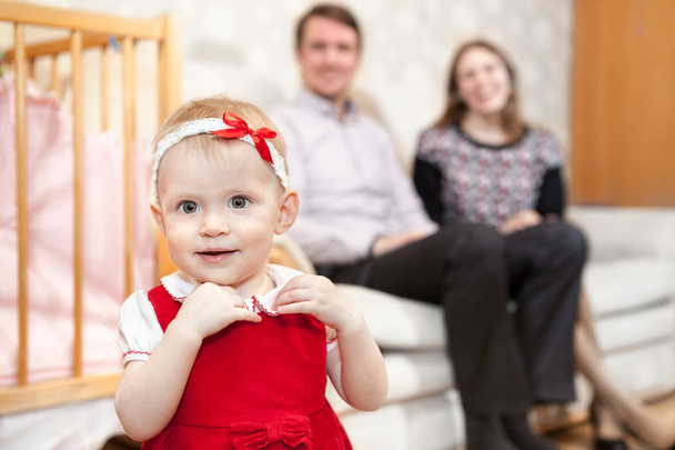 One year old Caucasian baby in red dress with parents are on background - Photo, Image