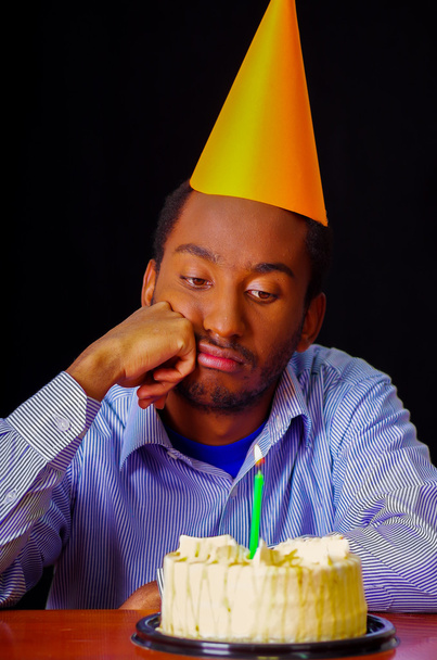 Sad man wearing blue shirt and hat sitting by table with cake in front, single candle burning, looking bored depressed, celebrating alone concept - Foto, afbeelding