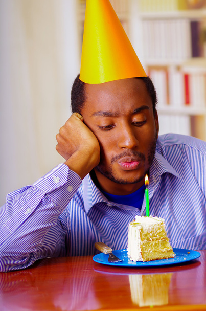 Sad man wearing blue shirt and hat sitting by table with piece of cake in front, looking depressed, celebrating alone concept - Photo, Image