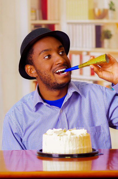 Charmante man dragen blauw shirt en hoed zittend op tafel met verjaardagstaart aan de voorkant, blazen partij hoorn vieren alleen glimlachend - Foto, afbeelding