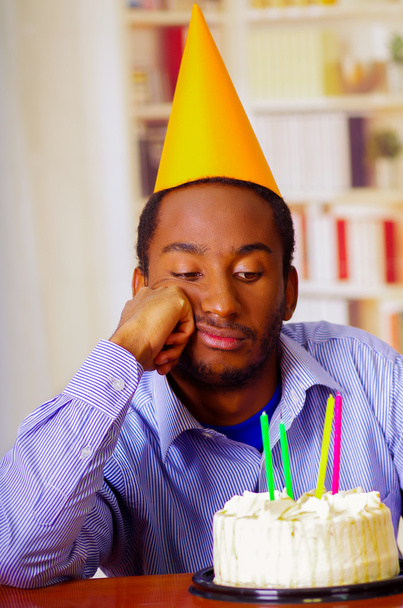 Charming man wearing blue shirt and hat sitting by table with birthday cake in front, looking sad depressed celebrating alone - Foto, afbeelding