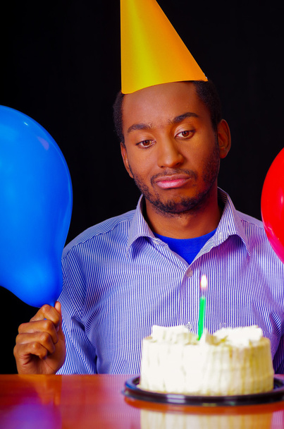 Good looking man wearing blue shirt and hat sitting by table with cake in front, single candle burning, holding balloons facing camera, celebrating alone concept - Photo, Image