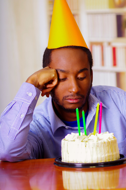 Charming man wearing blue shirt and hat sitting by table with birthday cake in front, looking sad depressed celebrating alone - Foto, immagini