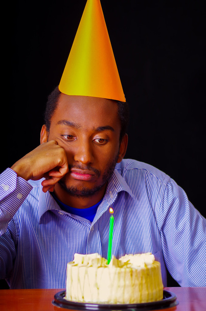Сумний чоловік у блакитній сорочці і капелюсі сидить за столом з тортами спереду, одна свічка горить, виглядає нудною в депресії, відзначаючи лише концепцію
 - Фото, зображення