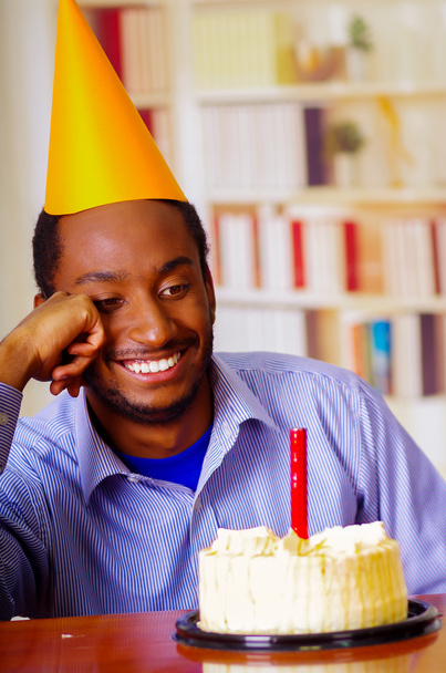 Hyvännäköinen mies, jolla on sininen paita ja hattu ja joka istuu pöydän ääressä kakku edessä, yksi kynttilä palamassa, skeptisesti hymyilevä, juhlii yksin.
 - Valokuva, kuva