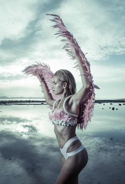 Romantic young beauty as an angel on the beach. Side view of blonde pretty female woman wearing pink wings, feather bra and bikini bottom standing in the sea over cloudy sky background  - Photo, Image