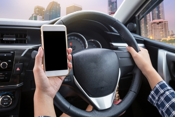 Fiatal nő vezető használ érintőképernyős okostelefon és kézi kormány egy autó város alkonyat háttér - Fotó, kép