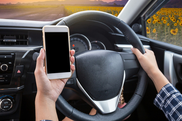 Mladá žena řidič pomocí dotykové obrazovky smartphone a ruka drží volant v autě se slunečnice pole za soumraku čas na pozadí - Fotografie, Obrázek