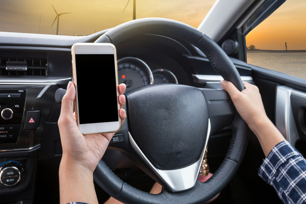 Mladá žena řidič pomocí dotykové obrazovky smartphone a ruka drží volant v autě s větrnou turbínou při západu slunce pozadí - Fotografie, Obrázek
