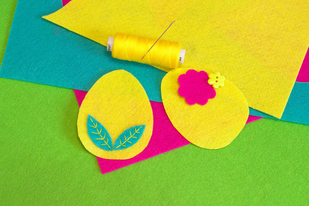 Пасхальний яєчний орнамент, жовта нитка, голка, кнопка, барвисті фетрові листи на зеленому тлі. Як зшити великодній яєчний декор. Домашній декор. Швейні вироби. Крок
 - Фото, зображення