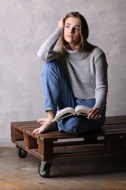 Girl with a book sitting on wooden board. Gray background - 写真・画像