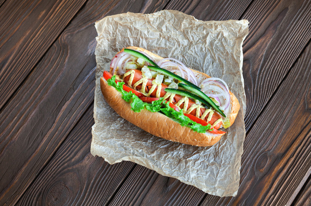 Big tasty hot dog with sausage, vegetables isousom on parchment on the wooden background. hot dogs to gourmet. - Фото, зображення