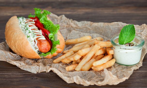 Big tasty hot dog with sausage and vegetables with sauce and French fries on the parchment on the wooden background. hot dogs to gourmet. - Fotó, kép