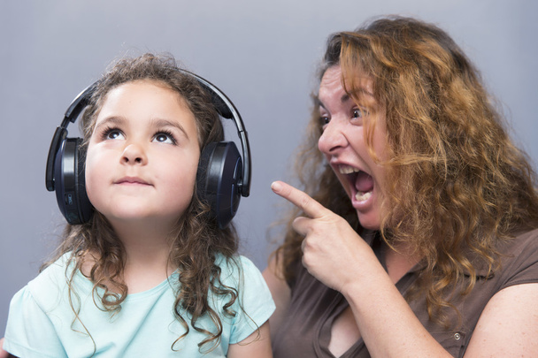 Lovely girl in headphones looking up while her mother screaming with index finger - Foto, Imagen