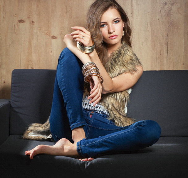Portrait of elegant woman sitting on black sofa wearing a blue jeans and fur vest - Fotó, kép