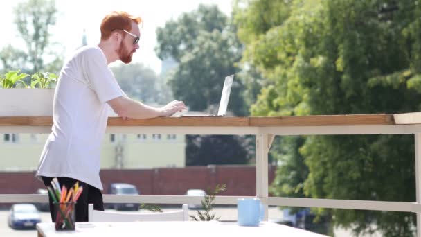 Молодой человек работает на ноутбуке, печатает, стоит на балконе на открытом воздухе
 - Кадры, видео
