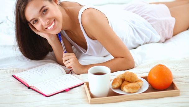 Young beautiful woman sitting in bed and writing in a copybook - 写真・画像